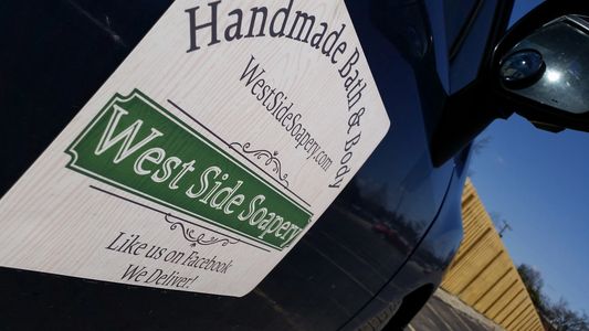 A blue car with a West Side Soapery magnetic sign attached to the side of it.