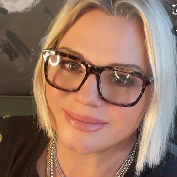 Woman with blonde hair and large tortoiseshell glasses smiling.