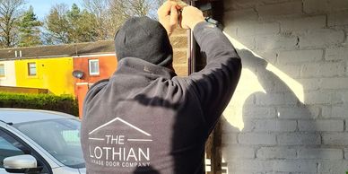 Man measuring a garage with the logo on back on top The Lothian Garage Door Company
