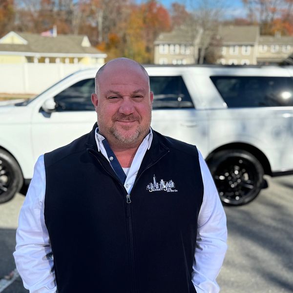 Smiling man in a black vest stands in front of a white SUV on a sunny day.