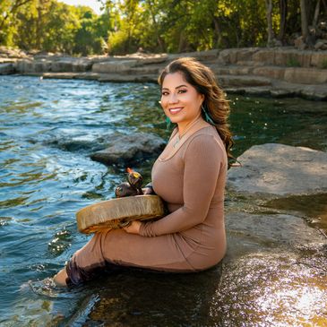 Woman in a brown dress sitting in the water holding a drum with carved birds.