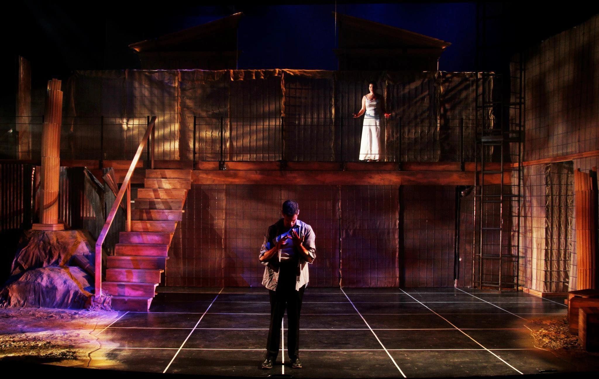 A dramatic stage scene with two actors, one illuminated in white above and one in shadow below.