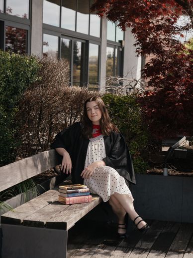 Young woman in a graduation gown sits on a bench with books outdoors.