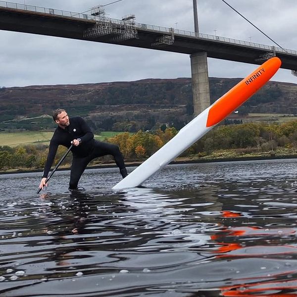 Man balancing on a large, orange and white paddleboard fin in water near a bridge.
