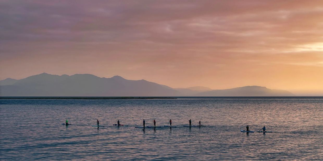People paddleboarding on calm water during a serene sunset with mountains in the background.