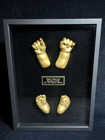 Gold coloured newborn baby hand and foot castings displayed in a black shadow box.