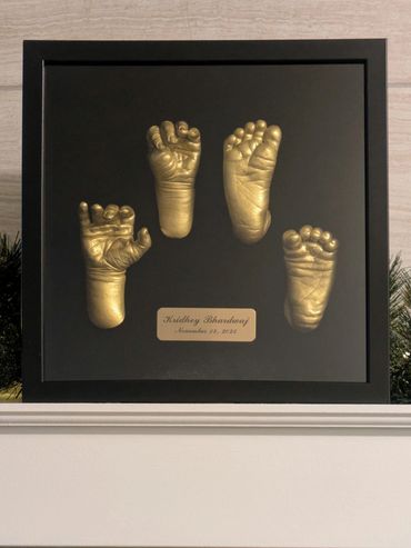 Gold coloured newborn baby hand and foot castings displayed in a black shadow box.