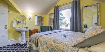 Cozy bedroom with yellow walls, patterned bedspread, a pedestal sink, natural lighting.