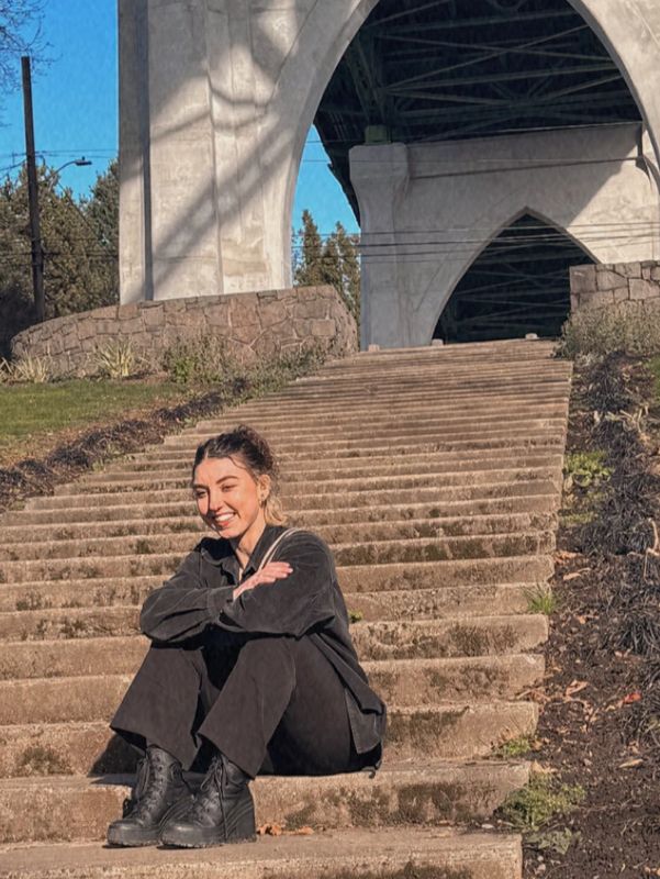 A woman sits on outdoor steps under a large concrete arch, smiling in the sunlight.