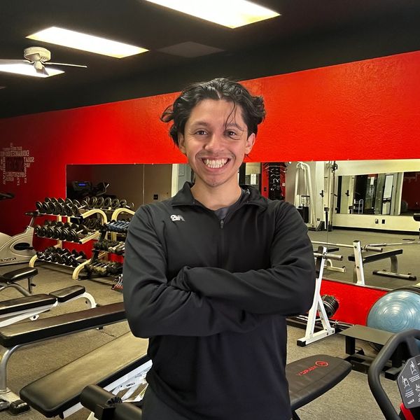 A smiling man stands confidently in a gym with workout equipment.