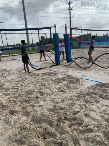 Group of people exercising with battle ropes on a sandy volleyball court.
