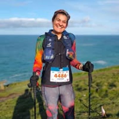 Smiling woman in colorful athletic wear with race number, holding trekking poles by the sea.