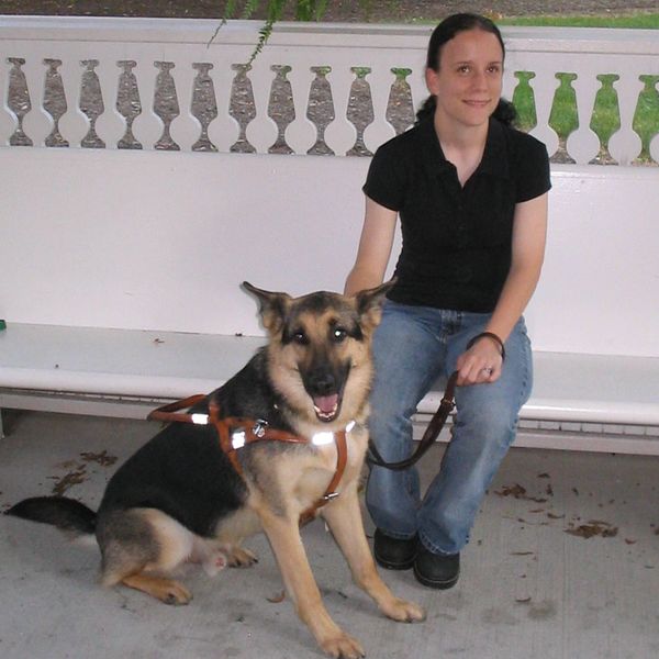 Photo of Heather sat on a bench with her Seeing Eye dog Rafe sat at her feet. Heather is wearing a b