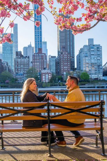 Couple sitting on bench under blooming cherry blossoms with cityscape.