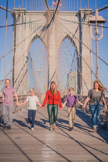 A family of five walking hand-in-hand across the iconic Brooklyn Bridge, captured during a vibrant outdoor family photoshoot in New York City. The bright sunlight and architectural backdrop highlight the strong family bond and the beauty of one of NYC’s most famous landmarks. Perfect for capturing timeless family memories in an iconic setting