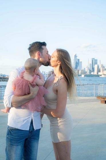 A family moment by the water: A man and a woman share an affectionate kiss while holding their baby with a city skyline in the background.