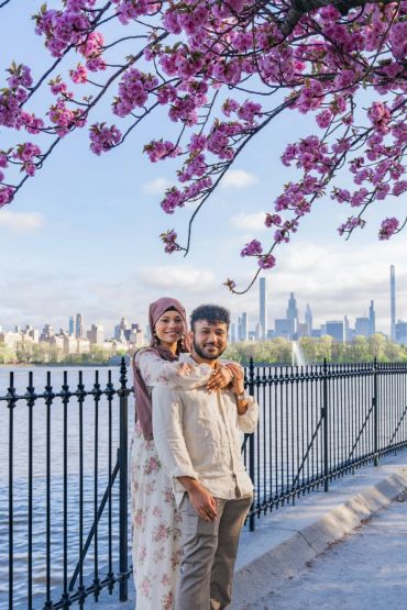 A couple embraces under pink blossoms by a city river.
