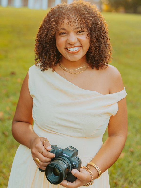 Woman in white dress holding a Canon camera, smiling outdoors.