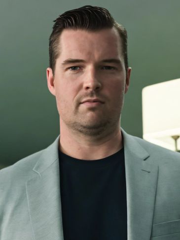 Man in a gray blazer and black shirt looking serious.