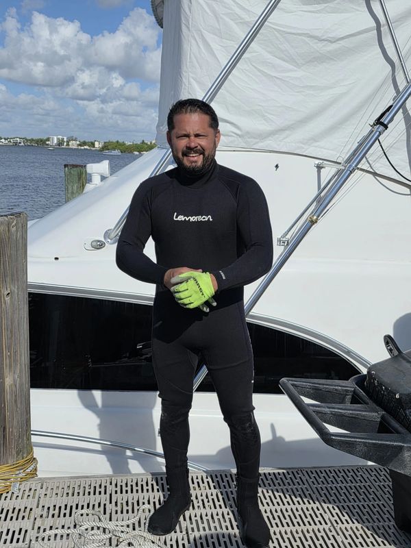 Man in black wetsuit and green gloves stands on boat dock by the water.