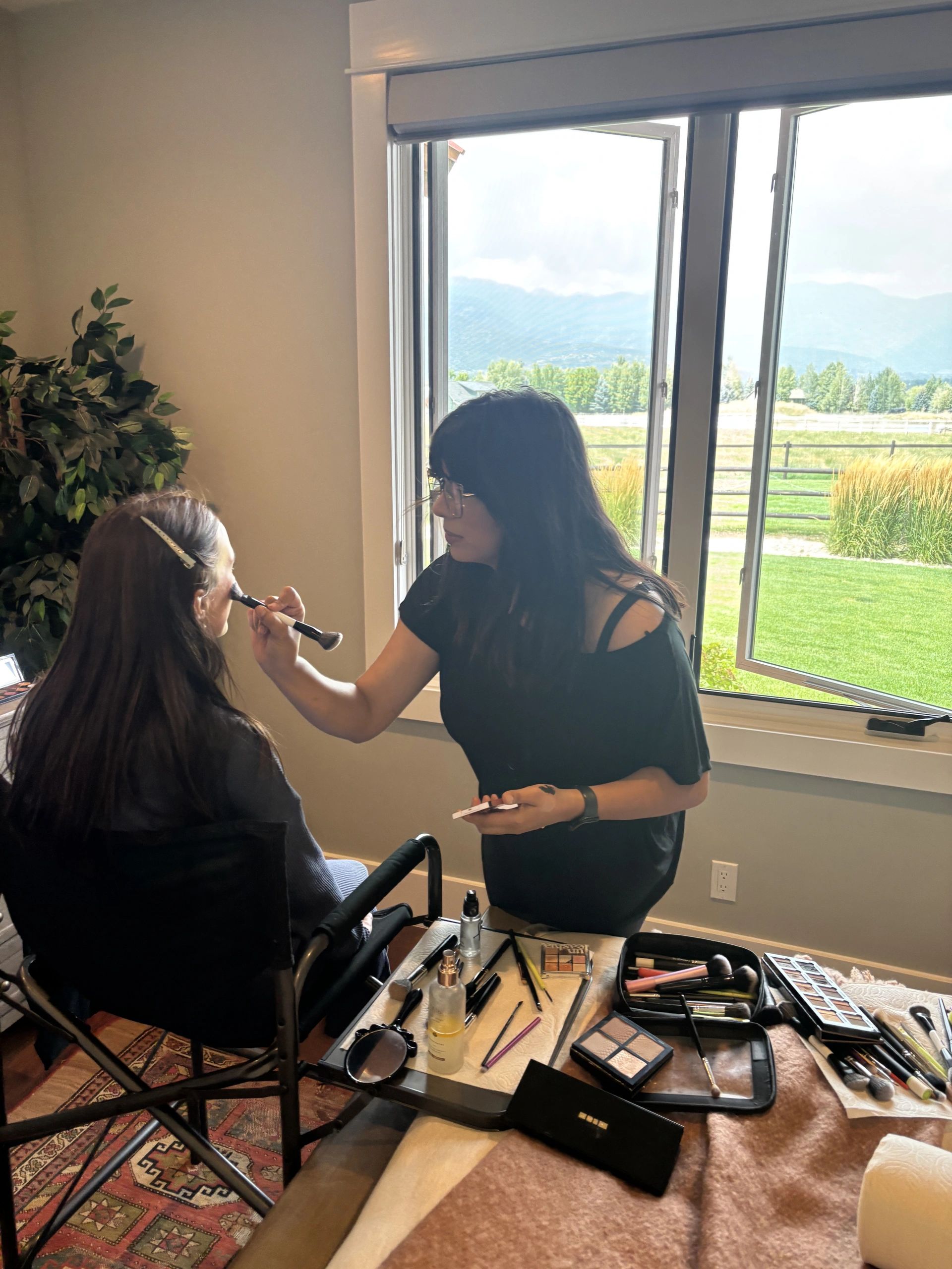 Makeup artist applying makeup to a seated woman near a window with scenic outdoor view.