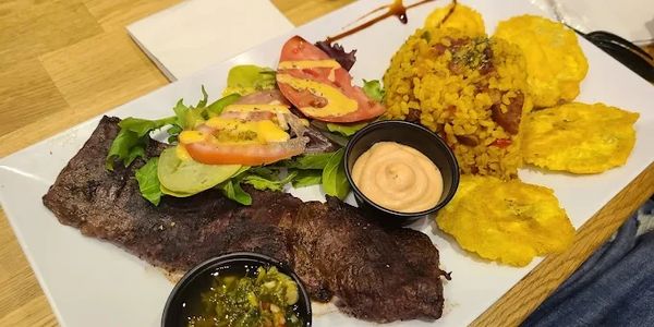 Grilled steak meal with salad, rice, tostones, and sauces on a white plate.