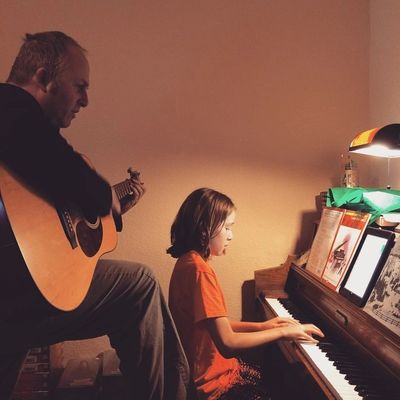 A man plays guitar while a girl plays piano in a cozy room.