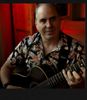 Man playing guitar in a dimly lit room with red background.