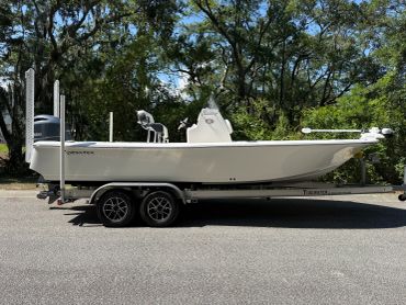 White boat on a trailer parked on a street with trees in the background.