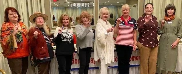 A group of eight women posing together indoors, each holding a small object.