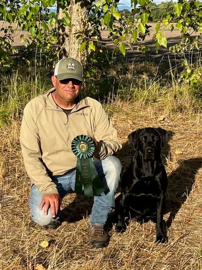Man kneeling with dog and award ribbon outdoors.