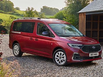 Red Peugeot van parked on gravel near a wooden structure in a green countryside.