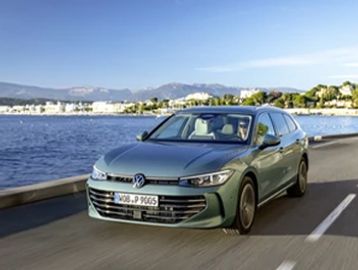 A Volkswagen sedan drives beside a waterfront road on a clear day.