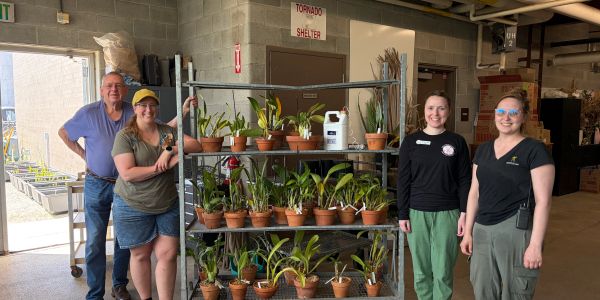 Four people smiling near a rack with potted plants.