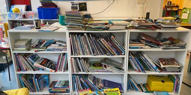 A cluttered classroom bookshelf filled with children's books and scattered piles.