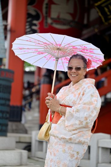 A smiling woman in a floral kimono holding a parasol at a traditional site.