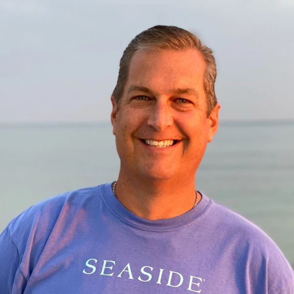 Smiling man wearing a blue 'SEASIDE' shirt at the beach during sunset.