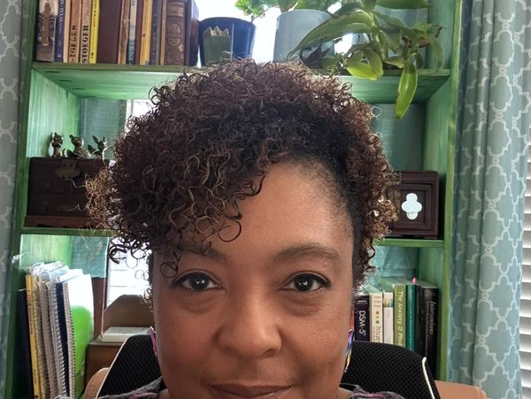 Woman with curly hair smiling in a cozy home office surrounded by books and plants.