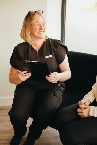 A smiling woman in black scrubs holding a clipboard, sitting and engaging in conversation.