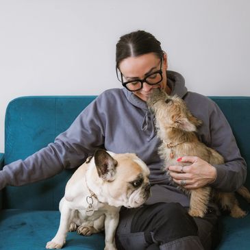 Woman on couch with two affectionate dogs, one licking her face.