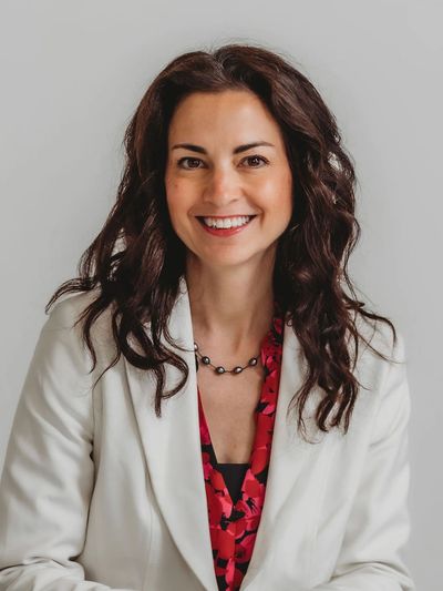 Smiling woman with curly hair in a white blazer and red floral blouse.