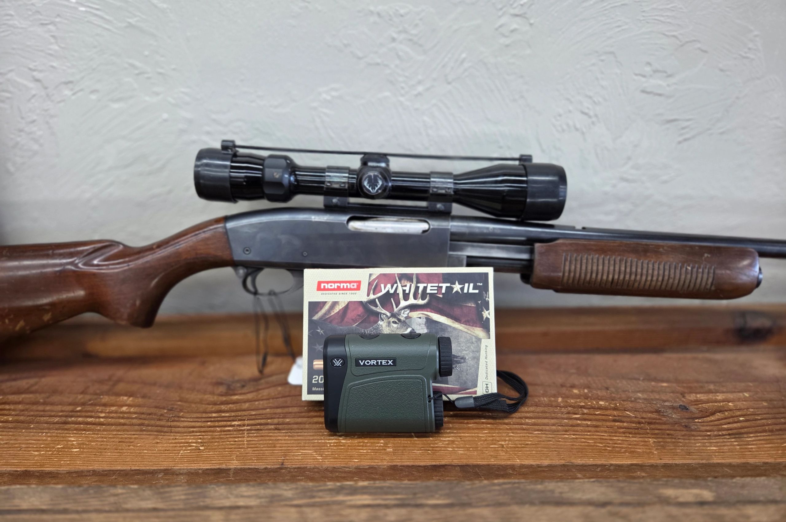 A rifle with a scope and a Vortex rangefinder on a wooden surface.