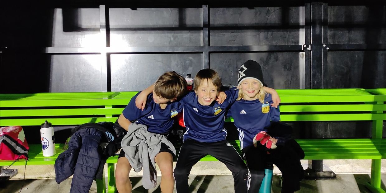 Three kids sitting on a green bench in matching sportswear.