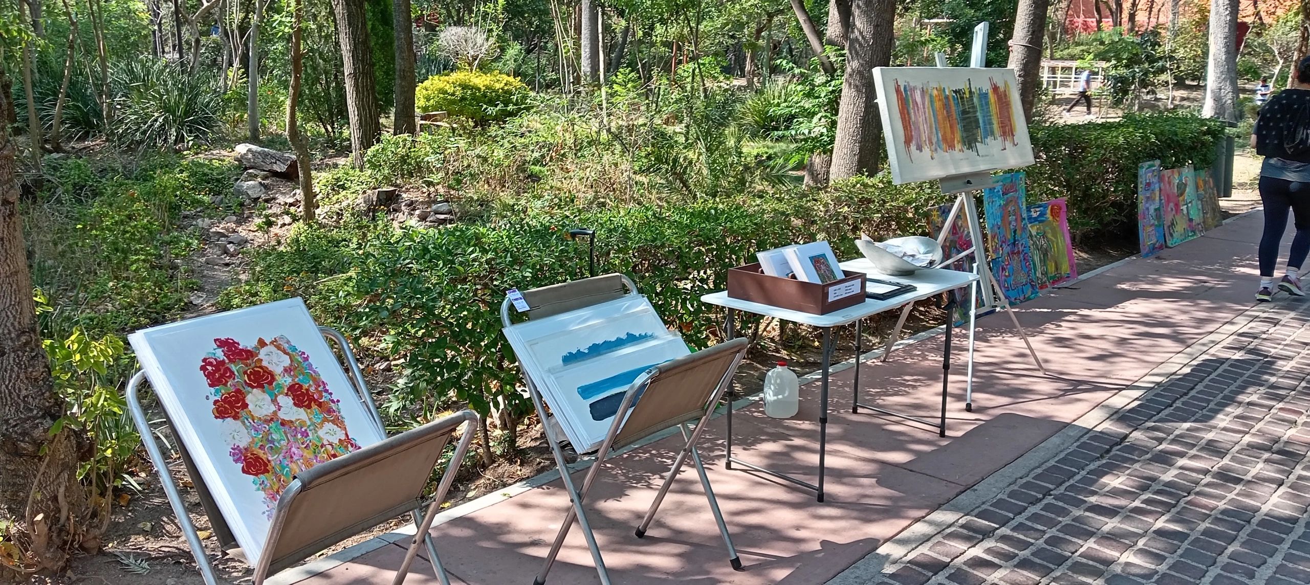 Outdoor art display with colorful paintings on chairs and an easel along a shaded path.