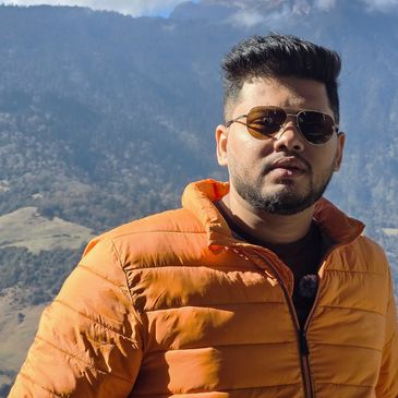 Man wearing an orange jacket and sunglasses with mountains in the background.