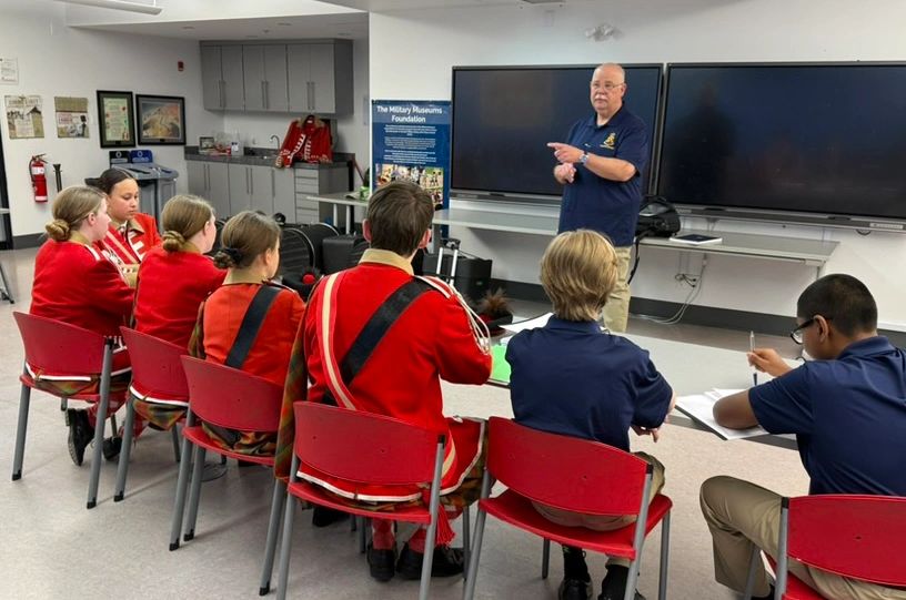 A group listens to a speaker in a classroom setting, some in red uniforms.