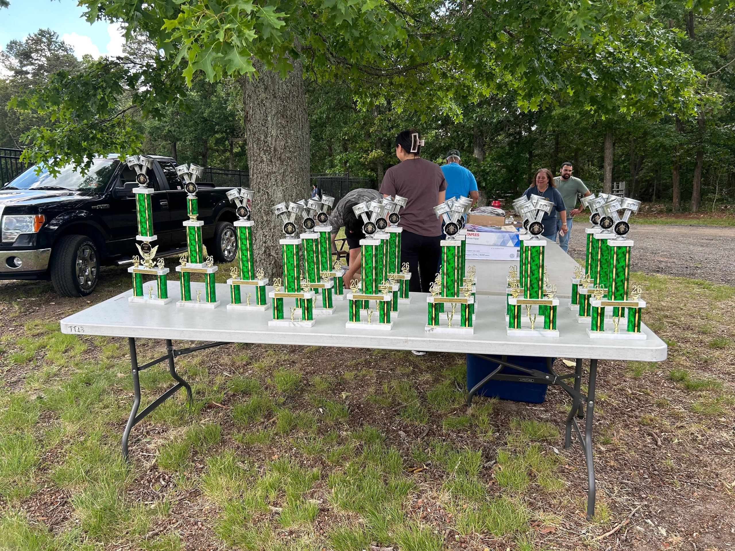 A table filled with green trophies outdoors with people and a truck in the background.