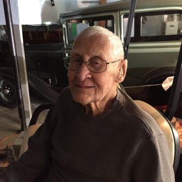 Elderly man smiling while seated indoors near a window.