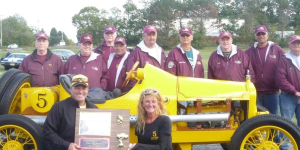 A group poses with a vintage yellow race car and a trophy outdoors.
