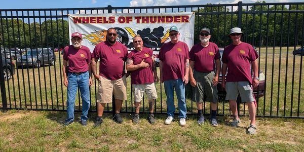 Six men in matching maroon shirts pose in front of a Wheels of Thunder banner outdoors.